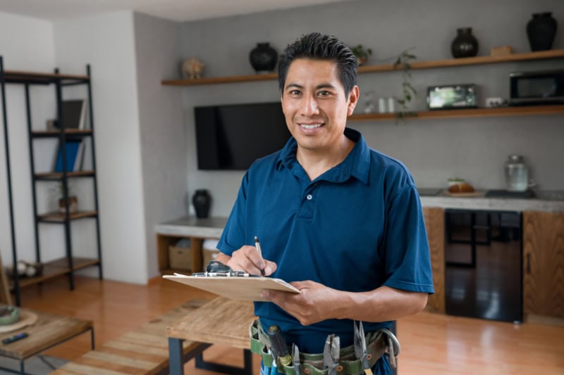 What Is a Plumbing Inspection? Happy Latin American electrician working at a house and looking at the camera smiling while writing on a clipboard - home improvement concepts.